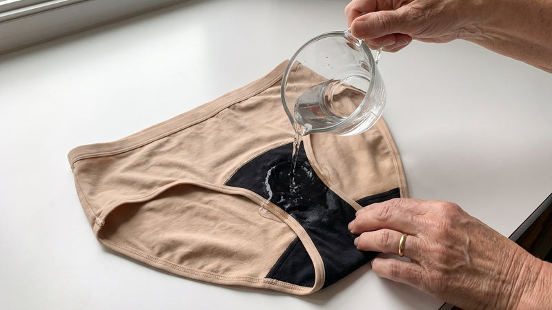 An older woman pours water from a glass measuring cup onto the black sewn-in core of a pair of Haven underwear on a kitchen counter